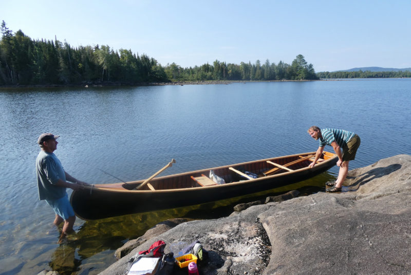 Two men hold a canoe near a rocky shore.