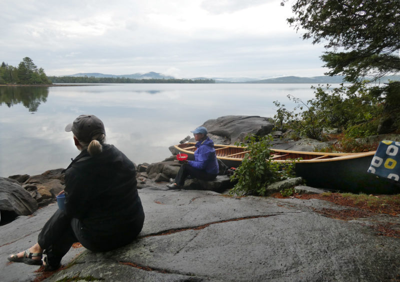 Two women eat breakfast on a rocky shore with a view of a lake.