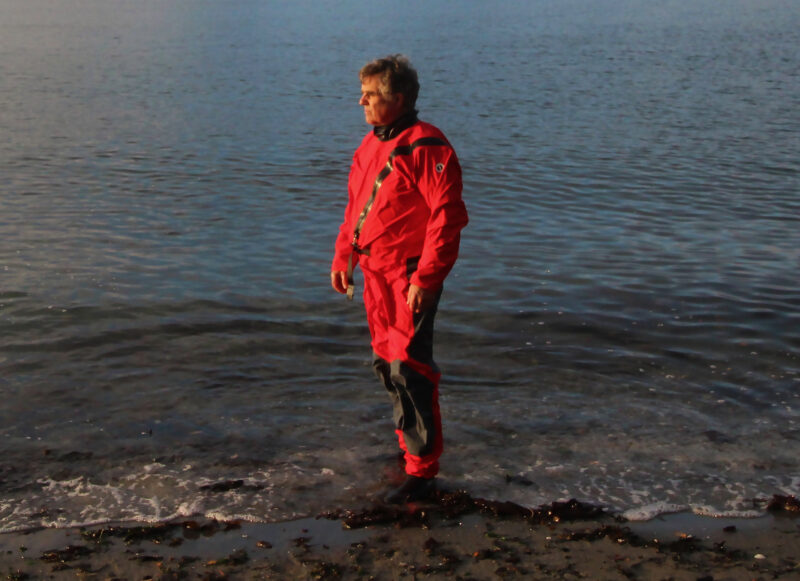 A man standing at the water's edge in a red Mustang Quadra drysuit