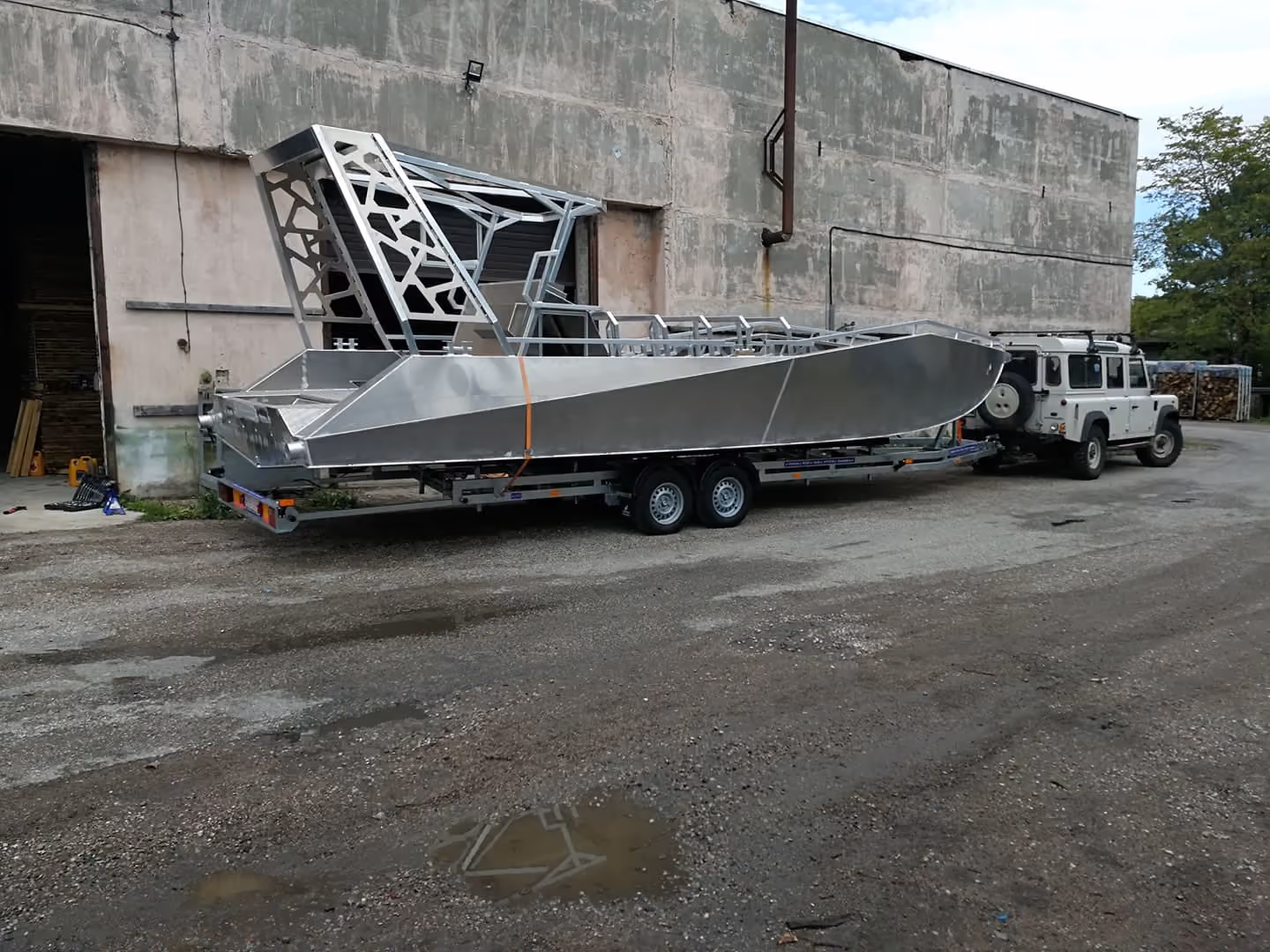 A bare, unpainted LR9 Safari passenger boat hull strapped to a twin-axle road trailer behind a vehicle, the aluminium wheelhouse frame welded up but not yet plated