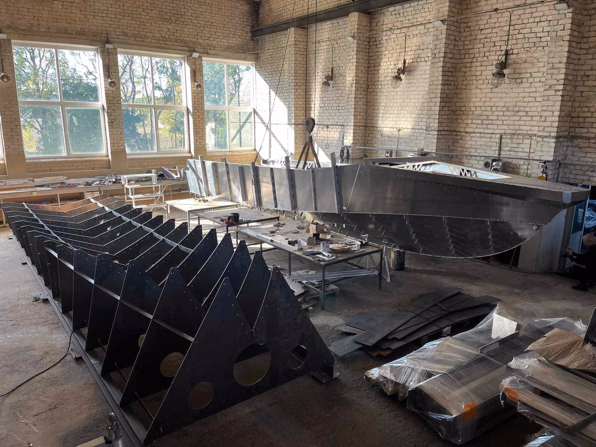 An LR9 Safari passenger boat hull under construction, the plate sides already welded up and a row of cut aluminium frames standing on a jig in the foreground