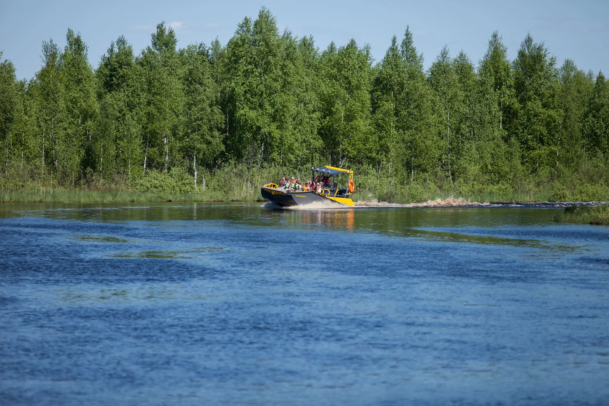 LR9 Safari passenger boat running a wooded river with a full load of passengers seated on the open forward deck and the skipper at the helm behind them